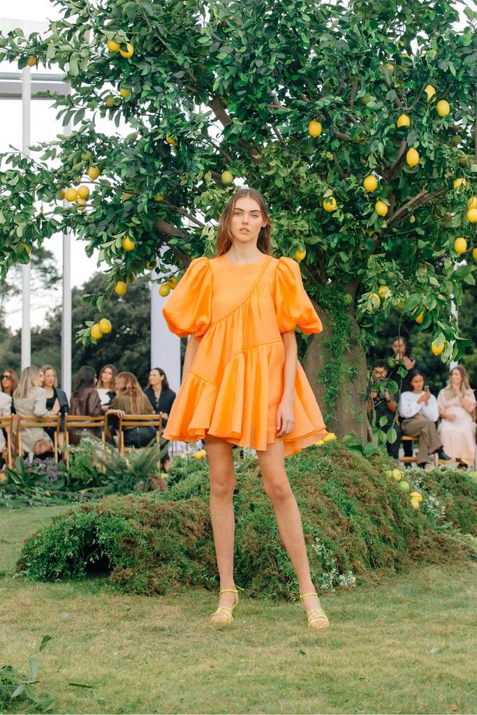 Model posing in AJE - RIVIERA ASYMMETRIC BRAIDED PUFF SLEEVE MINI DRESS - ORANGE in front of orange tree